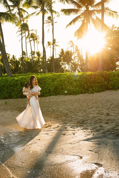 A woman wearing a long dress holds her baby as she walks along the beach with palm trees in the background.