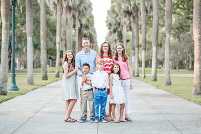 A family of 7 stands together in a large sidewalk lined with palm trees