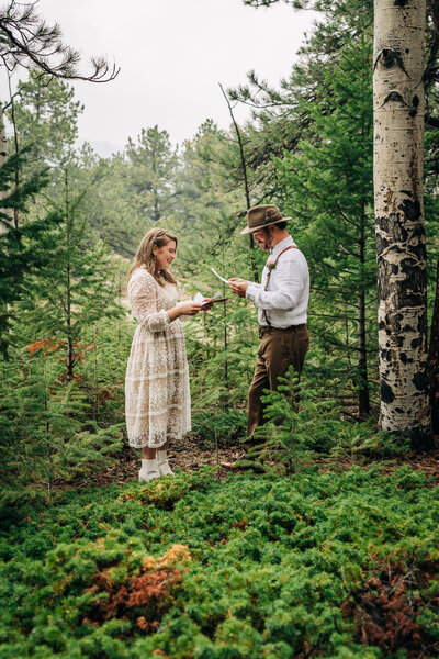 The couple is sharing vows in the forest together. They both have letters they're reading and there is trees and greenery surrounding them