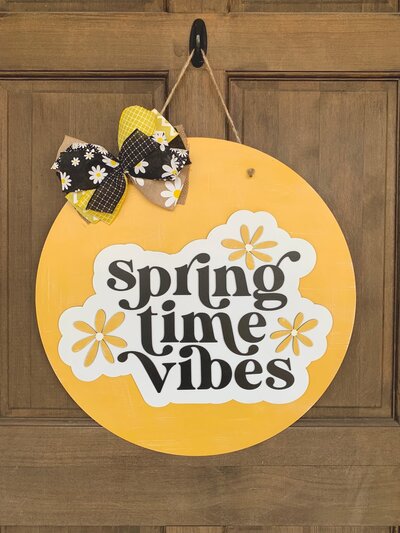 Yellow round door hanger with "spring time vibes" in black vintage font and yellow daisies