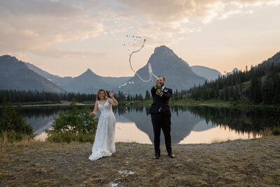 Bride and groom celebrate their Montana elopement by popping champagne in front of a mountain lake at sunset. The bride wears a lace wedding dress, and the groom, dressed in a black suit, sprays champagne into the air as the peaks of Glacier National Park reflect in the water behind them.