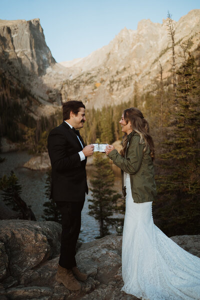 standing on a ledge overlooking an alpine lake. The bride and groom are smiling at each other and the bride is flipping her dress around so it blows in the wind.