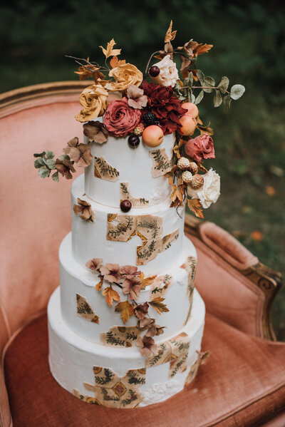 A four-tiered wedding cake with sugar flowers and fruit resting on a peach velvet vintage armchair