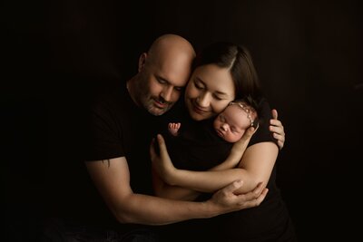 Mother and father lovingly embrace their newborn baby in a timeless studio portrait with rich shadows and warm connection, captured by Duehr Photography in Maple Grove,Mn