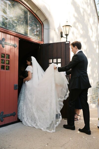 groom carries train for bride as the enter through red doors for wedding in minnesota taken by tom thornton photography
