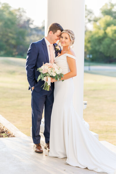 Bride and groom standing on a white-columned porch at golden hour, with the bride in an off-the-shoulder gown holding a blush and ivory bouquet. This elegant outdoor portrait was taken at a Denton wedding venue with soft natural light.