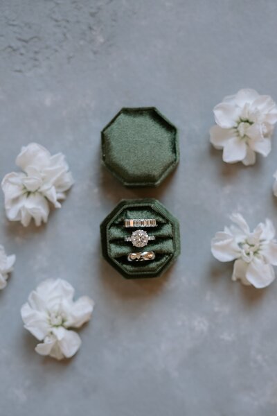 Wedding rings in a green velvet box laying with white florals on a flatlay surface.