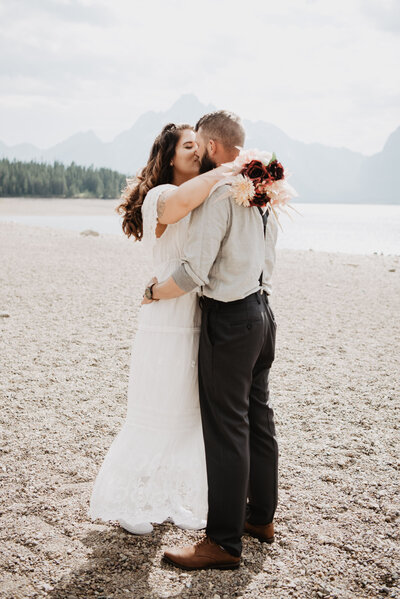 first kis between husband and wife at a jackson hole wedding  at Colter Bay Swim Beach with the bride holding her arms on her groom shoulder as she leans in