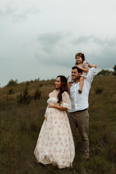 Pregnant woman smiling while her husband holds toddler daughter on his shoulders