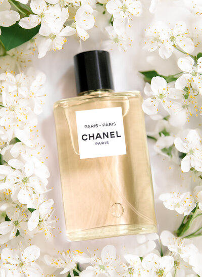 A bottle of Chanel Paris-Paris perfume is surrounded by delicate white flowers. The bottle has a simple, elegant design with a black cap and a white label. The scene conveys a sense of luxury and freshness.