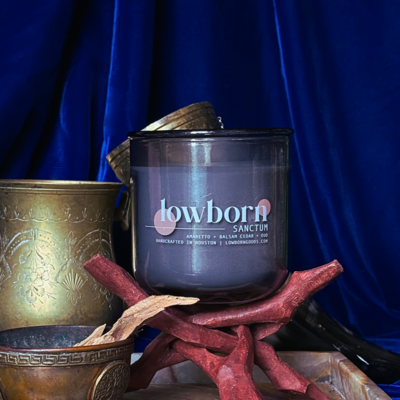 The homegrown candle, sitting on a wooden box. The candle is surrounded by vines, leaves, and greens, as well as a bunch of tomatoes. The image evokes a fresh herb and tomato garden on a sunshiny day.