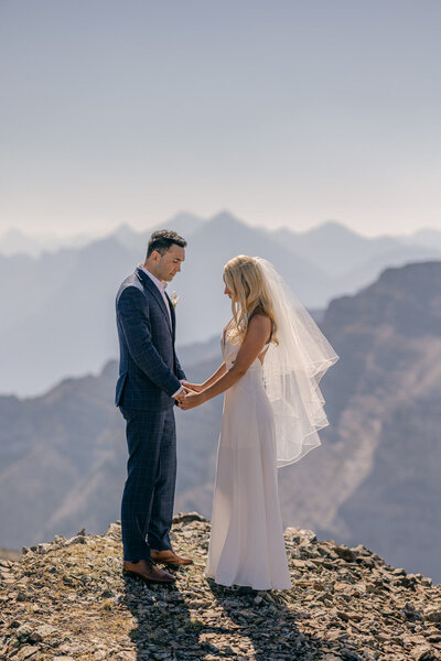 This serene portrait captures the bride and groom softly looking down at each other's hands, surrounded by stunning mountain views after their helicopter wedding elopement. The intimate and peaceful moment reflects their deep connection amidst the grandeur of their adventurous celebration.