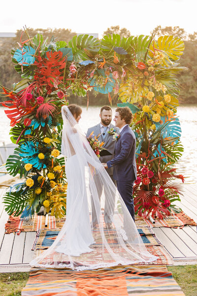 Colorful outdoor wedding ceremony by the water with a tropical floral arch, planned with bold, joyful flair in Austin, TX.