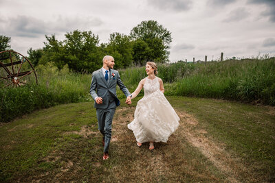 bride and groom run holding hands through a farm field