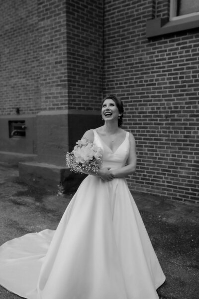 bride in wedding dress laughing holding floral bouquet standing in front of brick wall
