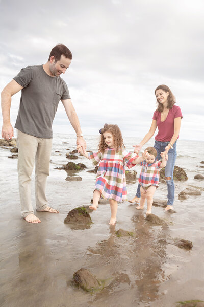 A dad, mom, and their two daughters happily walk barefoot along a rocky Connecticut beach. The parents are casually dressed, while the girls match in plaid dresses.