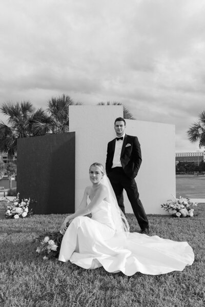 Modern bride in a sleek, minimalist white gown with a flowing train, seated gracefully against a geometric backdrop of neutral panels, surrounded by lush white floral arrangements. A sophisticated and editorial wedding portrait set against palm trees and an open landscape