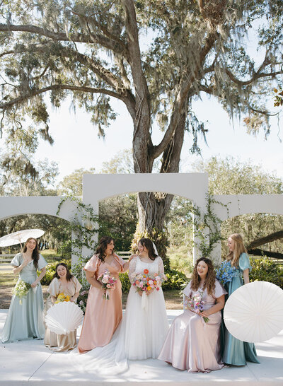 Bride and bridesmaids in pastel dresses posing under oak trees at Uliana Estate in Lakeland, Florida, with white parasols and garden arches in the background