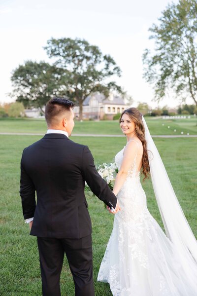 Bride and groom hold hands while walking across a lush green lawn, with the bride glancing back over her shoulder and smiling. She wears a lace wedding gown with a long veil, and the groom is dressed in a black tuxedo. A charming estate and large trees provide a romantic, scenic backdrop.








