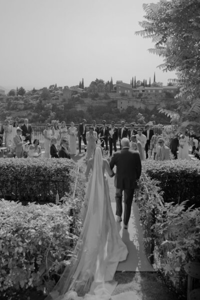Father of the Bride escorting his daughter during the Processional of a Gordes wedding overlooking the Luberon Valley. Guests are standing in a circular ceremony, surrounded by white and blue florals on all sides.
