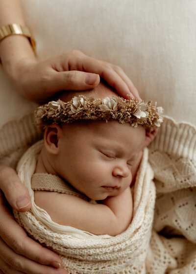 A close up of a relaxed sleeping baby wearing a flower crown  cradled by her mum's hands