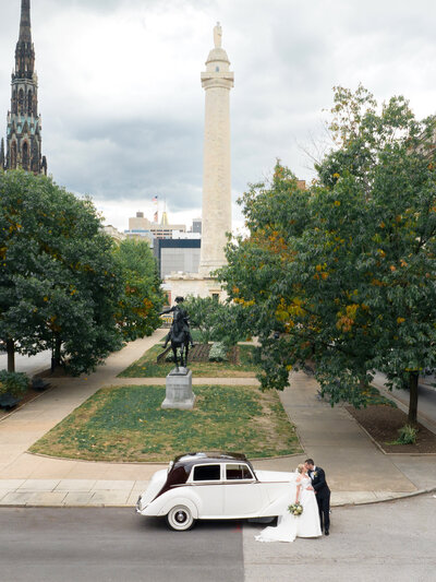 A couple in wedding attire kisses beside a classic white car on a city street. Behind them is a tall monument topped with a statue. Lush green trees line the scene on a cloudy day, adding a vibrant backdrop to the urban setting.