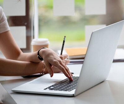 A woman uses a laptop with one hand while writing with the other