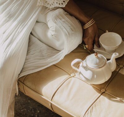 A soft tea moment unfolds with a teacup and teapot resting on a tan cushion, surrounded by white fabric and light.