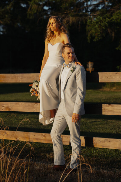 bride sitting on a fence, groom looking away at golden hour during wedding portraits in Comox by Latitude 49 Photography