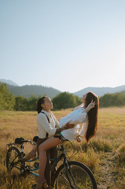 A candid lifestyle shoot of a couple riding a bicycle in a grass field. A woman is sitting on the seat of a bike while holding her girlfriend on the handlebars.