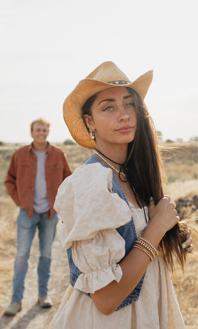 A close up of a woman wearing a cowboy hat. Standing behind her, slightly blurred is her boyfriend smiling at the camera.
