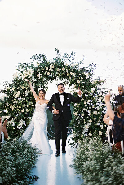 Bride and groom celebrating their wedding recessional beneath a floral arch with confetti — captured by Asia Pimentel Photography.