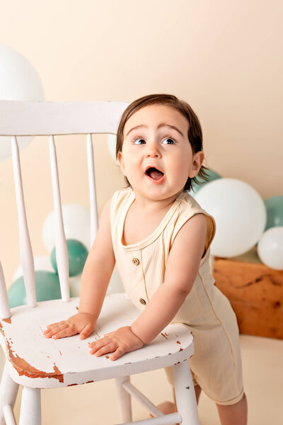 Toddle boy smiling with his mouth open standing against a white chair in front of a tan backdrop with green and white balloons.