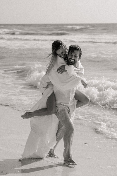 Black and white photo of a groom giving his bride a piggyback ride on the beach in Tulum, laughing in the surf.