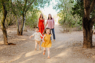 Family Photographer, two women hold hands as they walk behind their young children on a trail