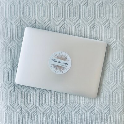 Image shows a closed silver laptop with a myffirmations affirmation card placed over the centre of it. The laptop is placed on a textured light blue background