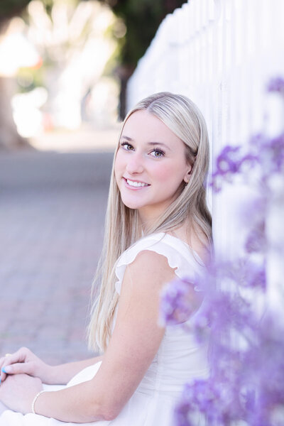 Young blonde woman with hazel eyes wearing a white dress sitting on a sidewalk and leaning against a white fence with purple flowers, looking to her left at the camera.