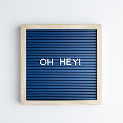 A wooden-framed letter board with a navy blue background and the phrase 'OH HEY!' in white letters, capturing a cheerful and welcoming vibe in a clean, modern design