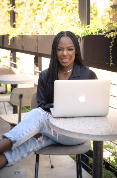 Rianne Nycole wearing a black top and jeans sitting a a table outside near a coffee bar. On the table in front her her is a silver macbook that's opened while she is smiling at the camera.