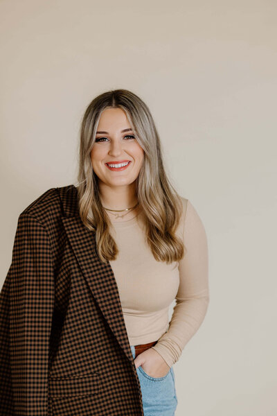 fun and laid back headshot of woman in neutral colors, jeans, and brown plaid blazer standing in front of neutral background