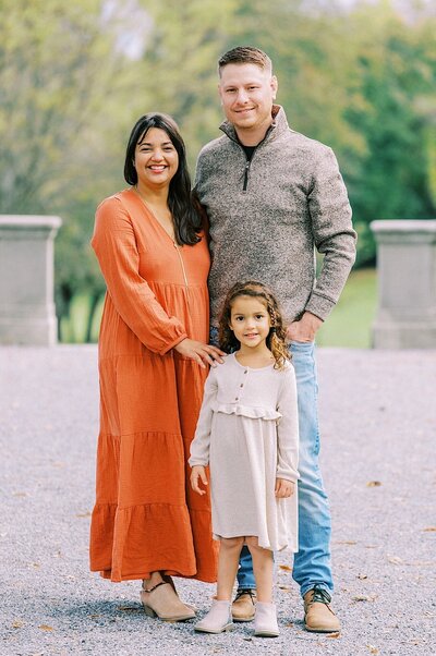 Family portrait with mom in orange dress and dad in gray sweater, with young girl