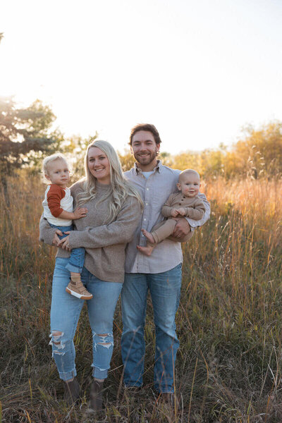 A happy family of four stands in a sunlit field, with both parents holding their young children close.