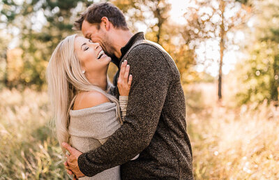 Husband embraces and kisses wife on the cheek, while she smiles during a photoshoot in the autumn forest