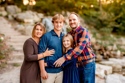 A blended family of four smiling at each other  while standing closely together as they enjoy their St. Louis family photo session.