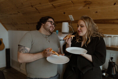 couple enjoying pizza and laughing together inside a cozy wood-paneled cabin kitchen during their intimate elopement getaway