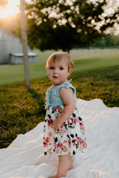 Toddler girl standing on blanket in blue and floral dress looking at camera on a farm with the sun shining behind her.