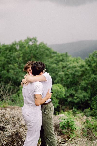 Cinematic wide shot of the couple embracing on the summit, with soft focus and natural light.
