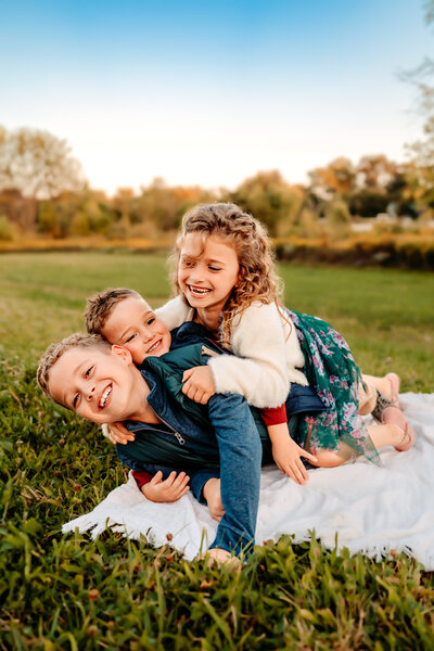 Siblings piled on top of each other smiling and laughing outside in the grass on a blanket