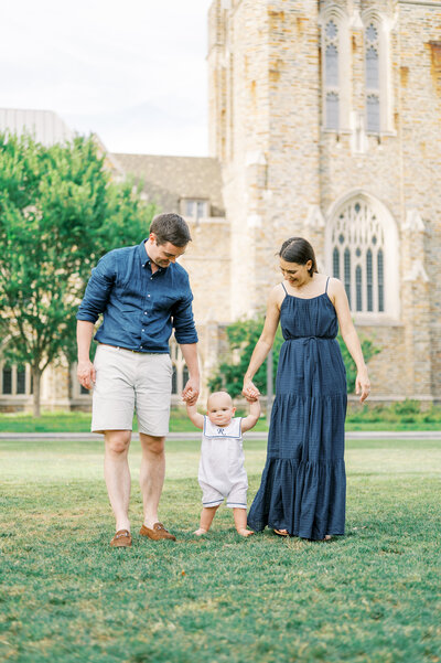 Mom & Dad hold hands of one year old boy during Summer family photo session outdoors