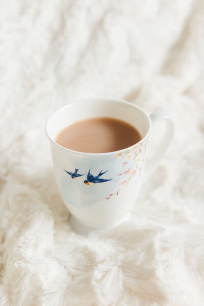 White china cup with blue birds pictured flying on it. Cup filled with tea sitting on a cream blanket.